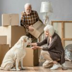 senior woman petting dog in new house while husband holding cardboard box